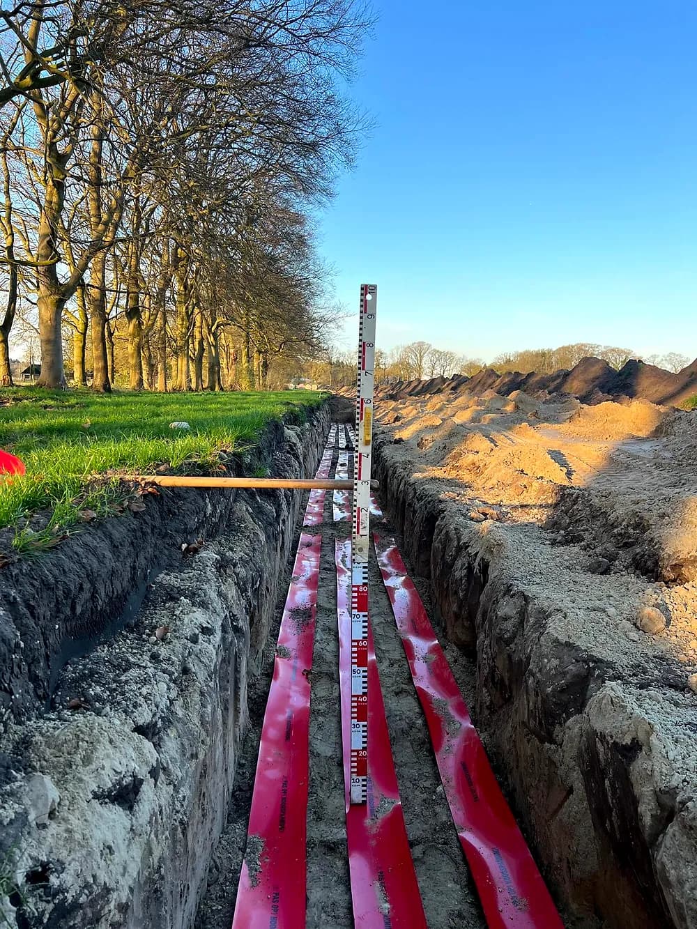 Rood afdekband in de sleuf met meetlat, bomenrij in de avondzon